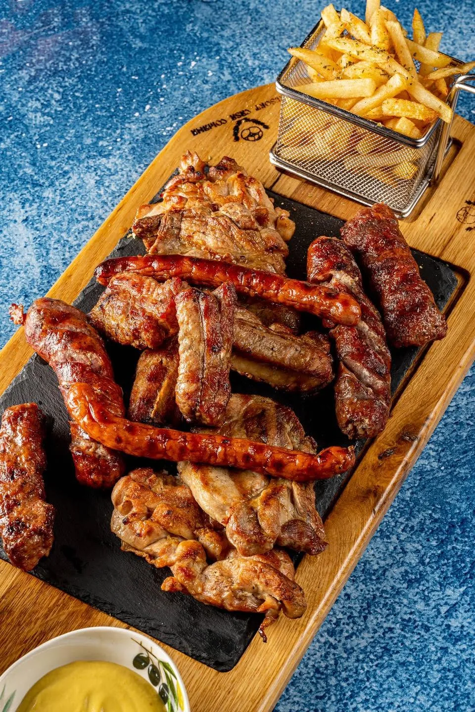 Platou cu cărnuri la grătar: cârnați, mici, ceafă și pulpe de pui, cu cartofi prăjiți și muștar, pe un fund de lemn